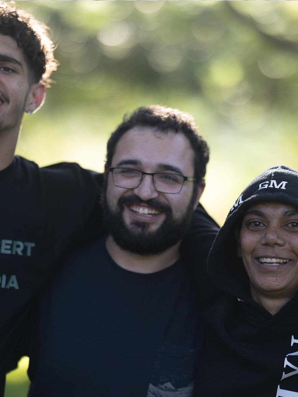 A close-up, joyful group photo of three people. The person in the center with glasses has their arms around the shoulders of the other two, and they are all smiling broadly.