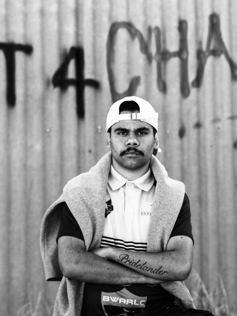 A black and white portrait of a young Indigenous man with a mustache, wearing a white baseball cap backwards and a polo shirt. A sweater is draped over his shoulders, and his arms are crossed, showing a forearm tattoo. He is standing in front of a corrugated iron wall with graffiti on it.