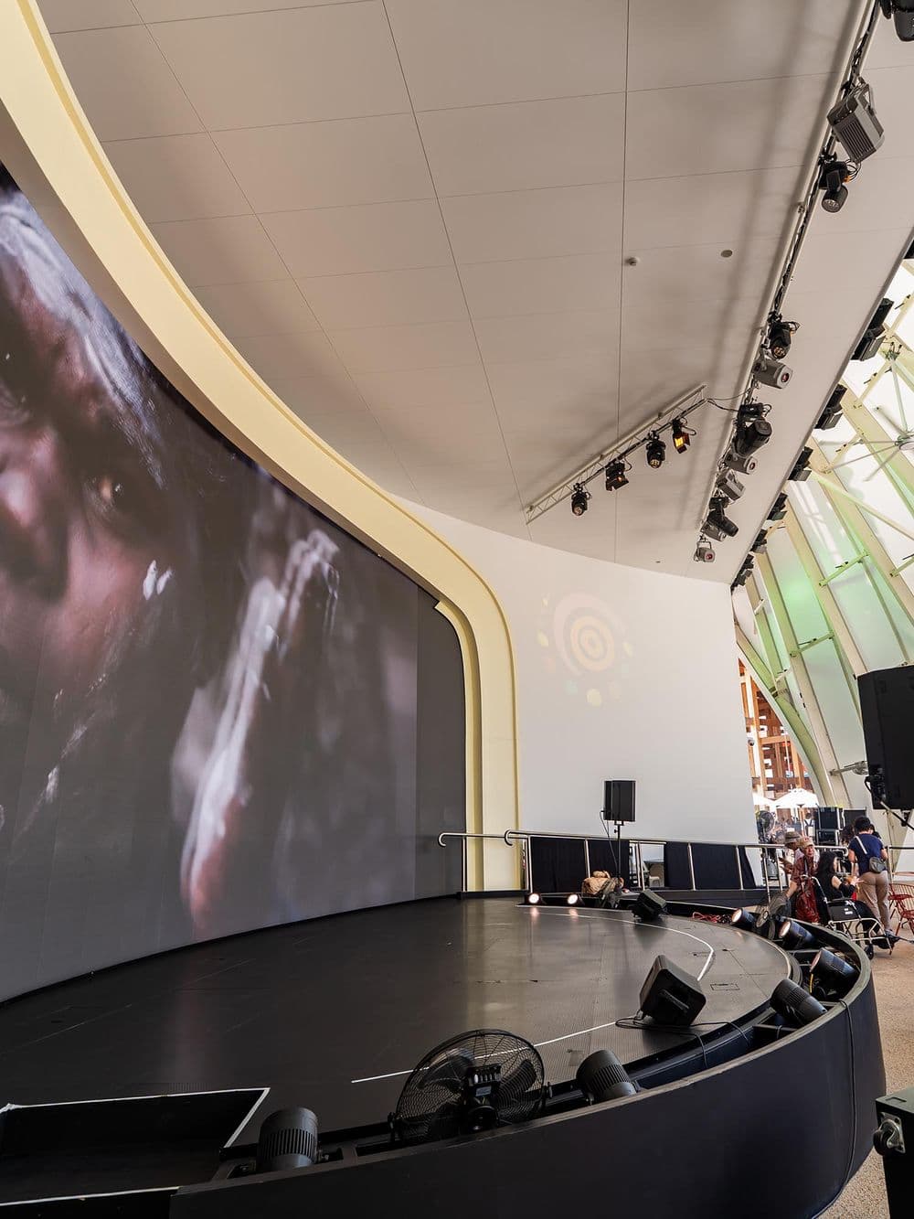 An indoor view of a modern stage with a large curved video screen on the left. The screen is displaying a close-up image of a person. The stage floor is black, and there are lights and speakers set up. The ceiling and walls have a white, modern design.