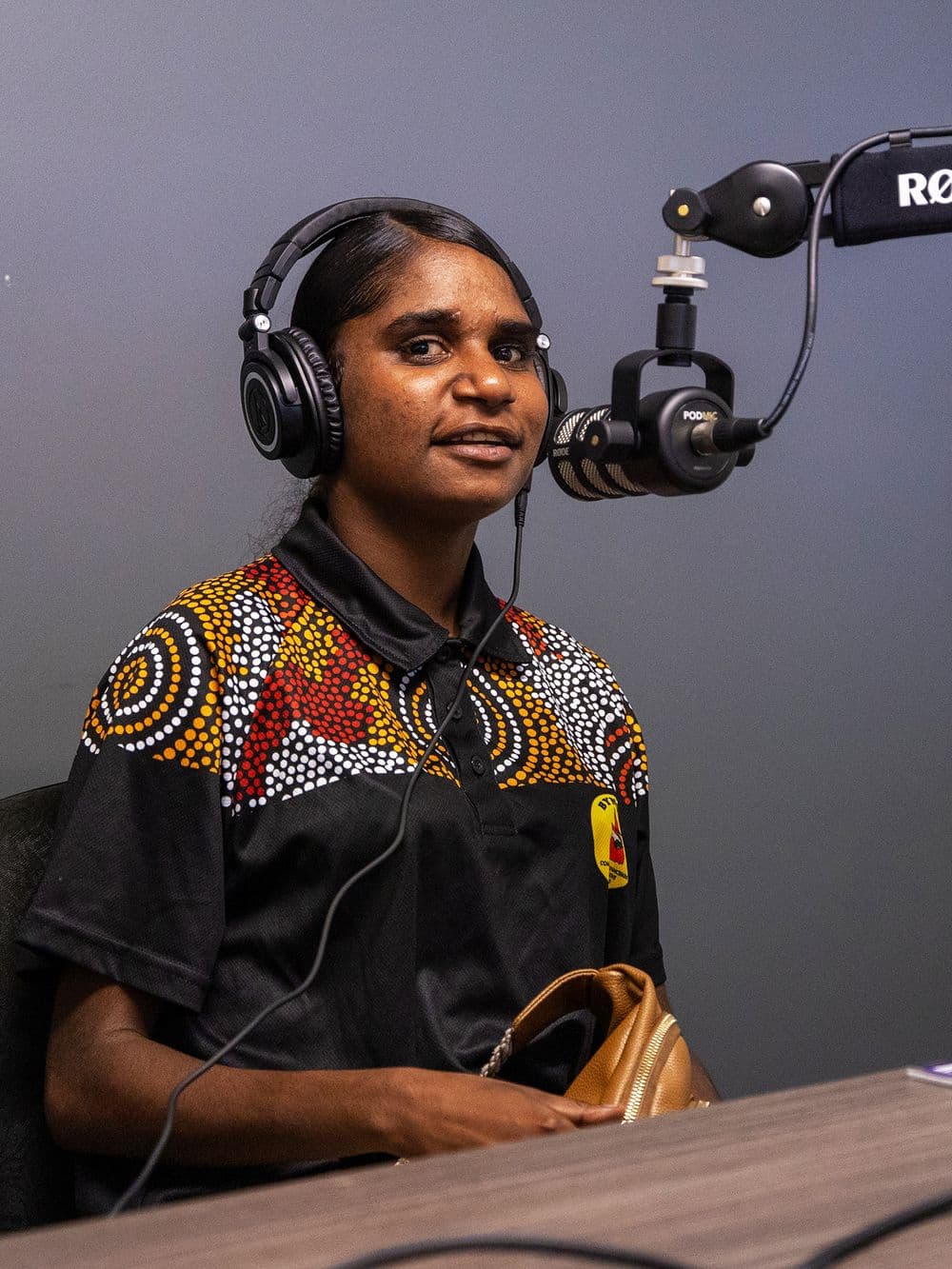 A young Indigenous woman with her hair pulled back is sitting at a desk in a recording studio. She is wearing headphones and an Aboriginal-designed polo shirt, and she is smiling while looking towards the camera. A microphone is positioned in front of her.