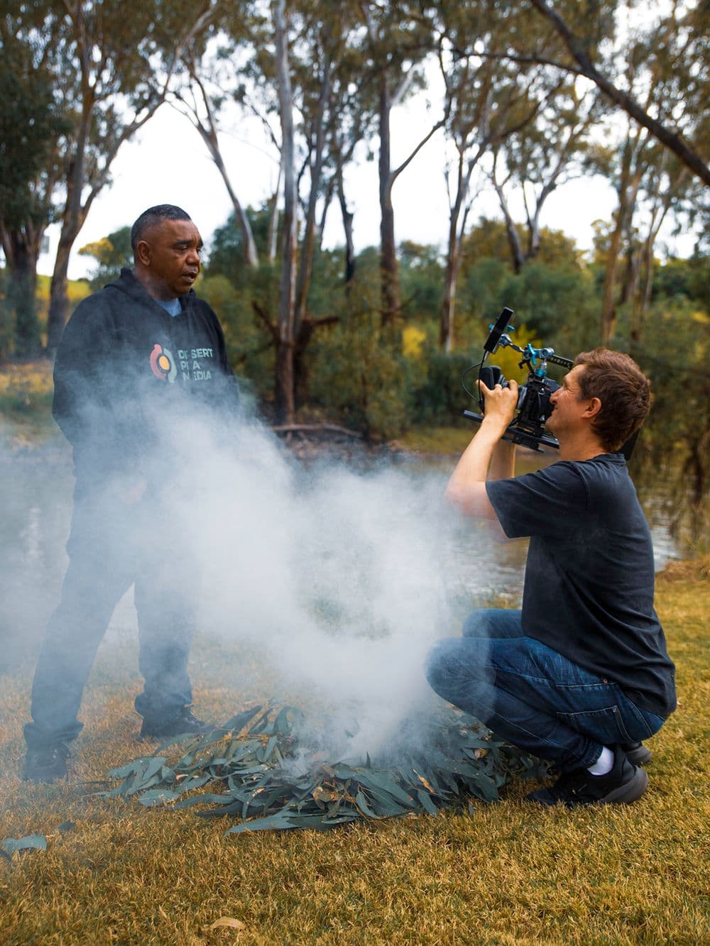 An Indigenous man in a black hoodie stands in a grassy area with trees in the background. He is watching a cloud of smoke rise from a traditional smoking ceremony. Another person, a cameraman, is kneeling and filming the scene.