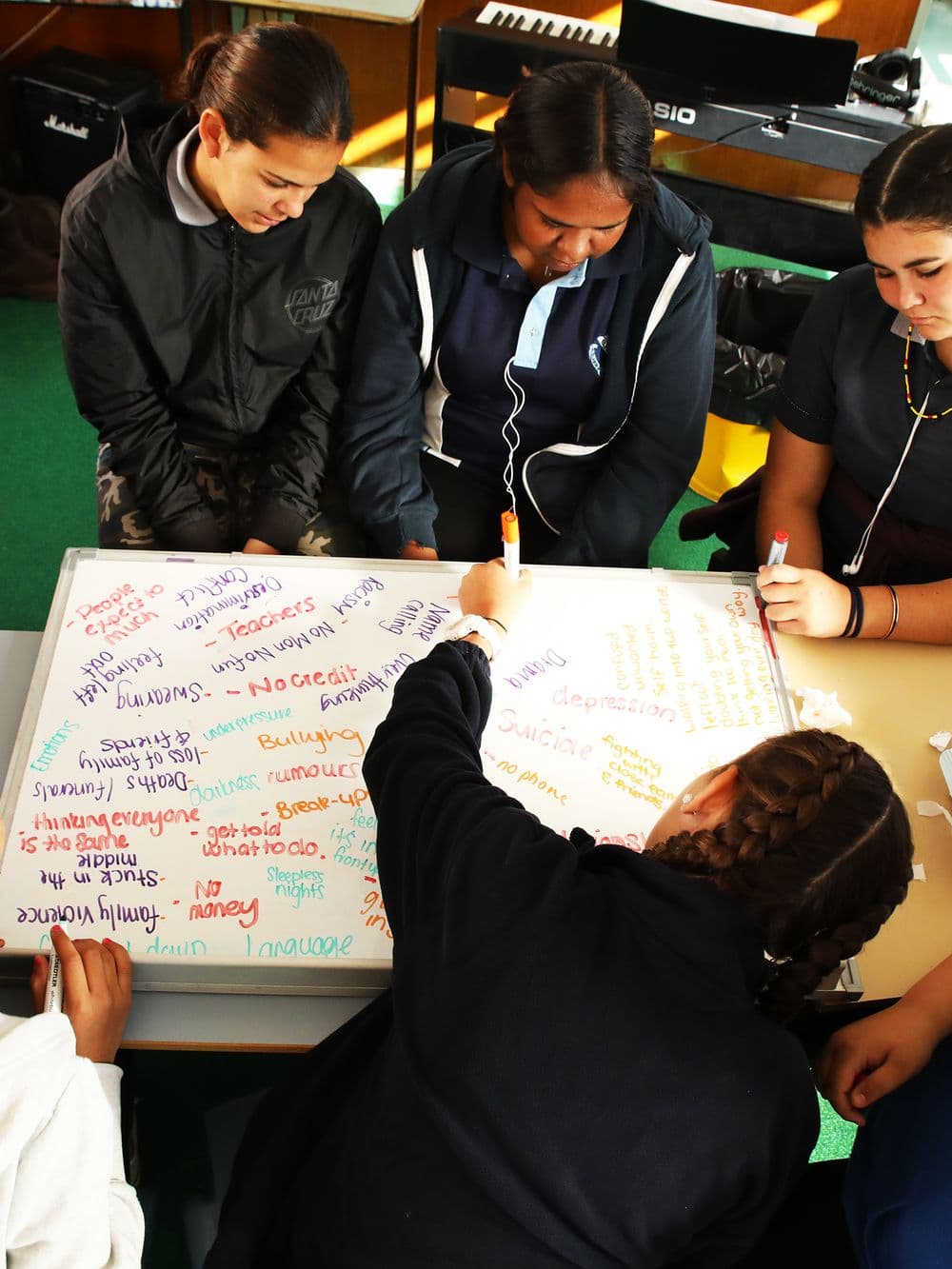 Four young people are gathered around a whiteboard on a table, writing on it with markers. The whiteboard is covered in handwritten words, and the young people appear to be collaborating on a project.