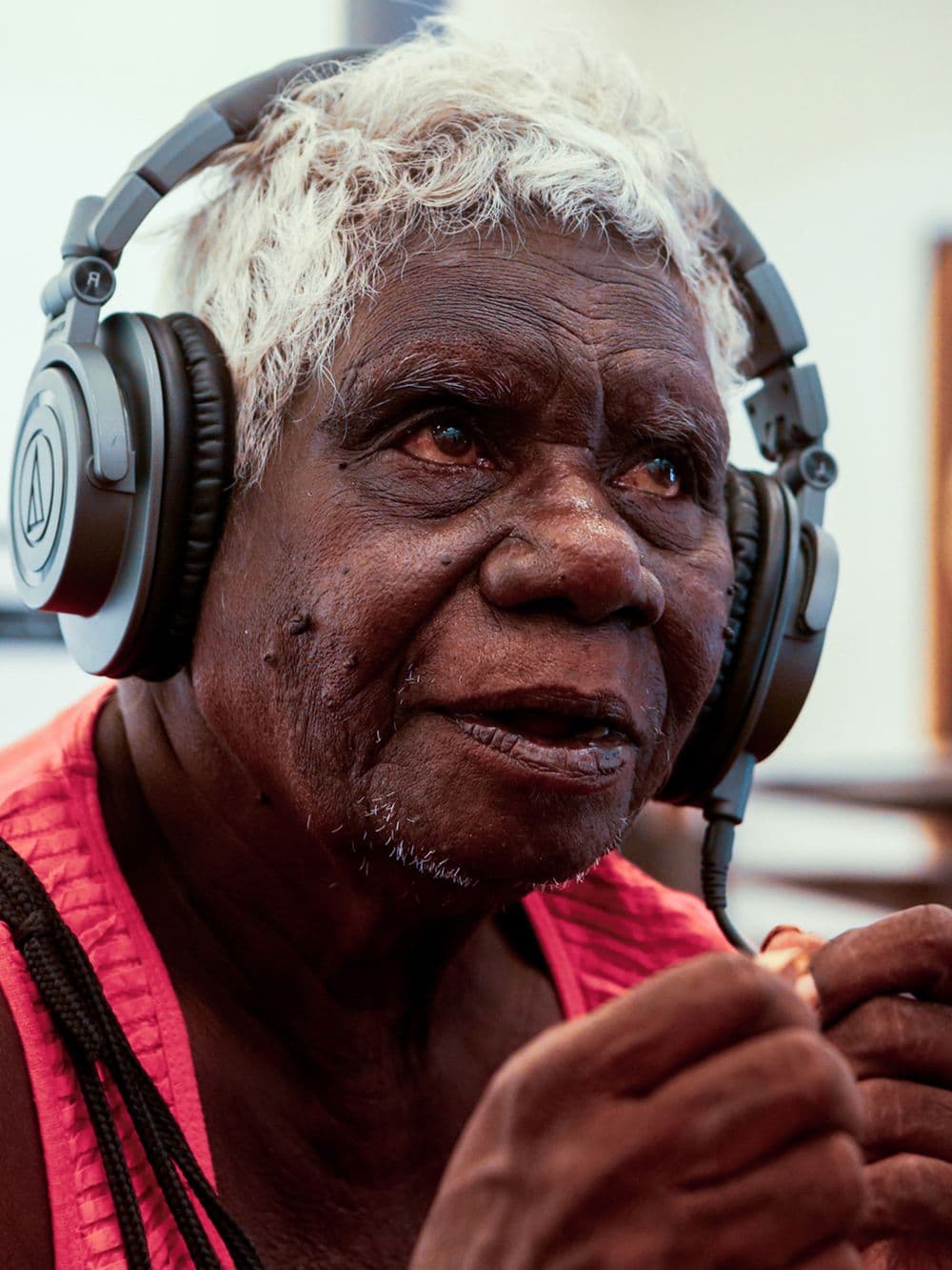 A close-up portrait of an elderly Indigenous person with white hair, wearing large headphones. They are looking up and to the side with a contemplative expression.