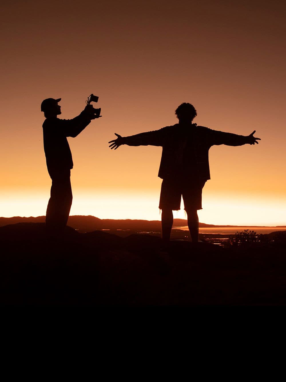 A silhouette of two people against a vibrant sunset sky. One person is on the left, holding up a camera as if taking a picture or filming. The person on the right has their arms spread wide, facing the scenic view.