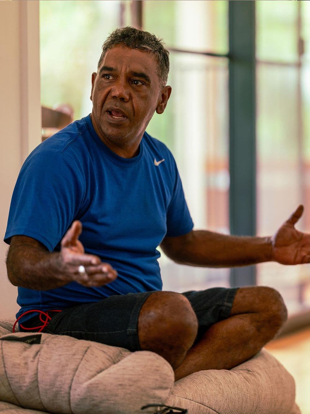 An Indigenous man in a blue t-shirt sits on a floor cushion, gesturing with his hands as he speaks. He is looking off to the side with a serious, engaged expression.