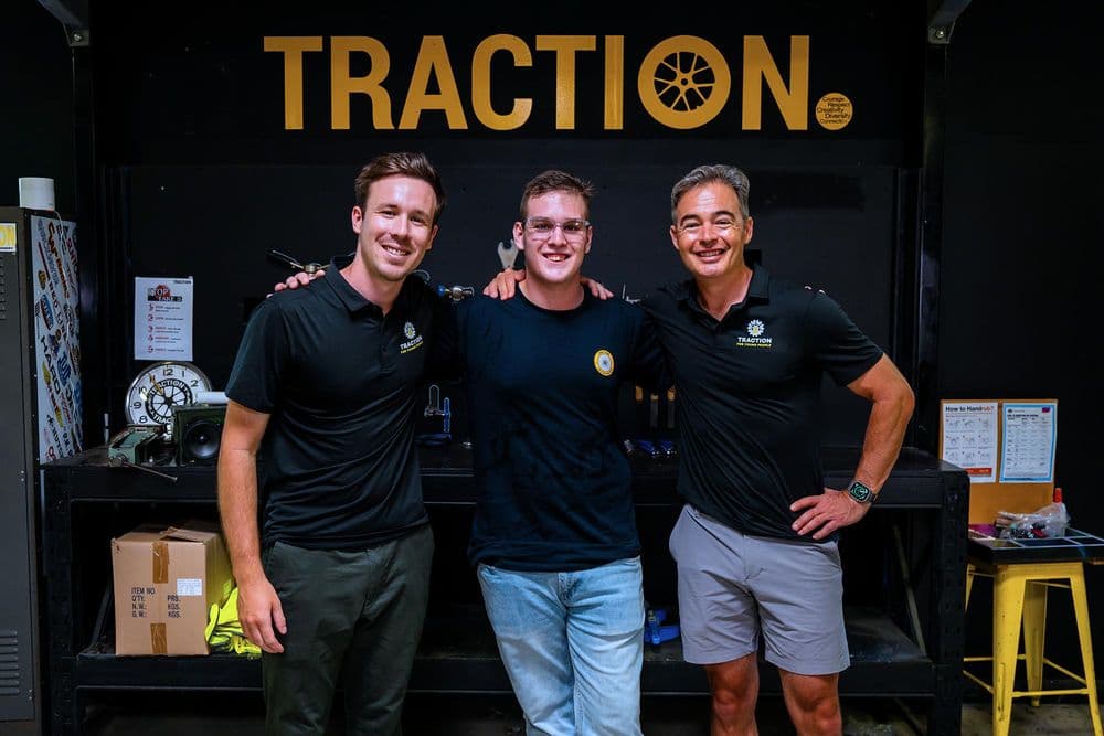 Three men are standing side-by-side and smiling in a workshop with a black wall that has a large yellow "TRACTION" logo on it. The man in the center, Riley, wears glasses and has his arms around the other two men, who are wearing black polo shirts with the Traction logo. A workbench is visible in the foreground.