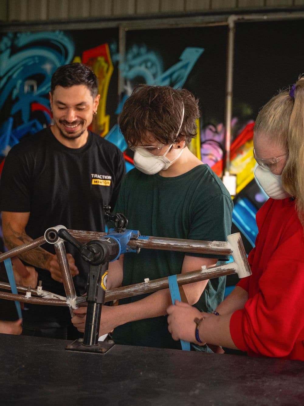 Three people are working on a bicycle frame mounted on a stand. Two young people are wearing white protective masks. A man wearing a black "Traction Mentor" shirt is standing behind them, smiling. Colorful graffiti is visible on the wall in the background.