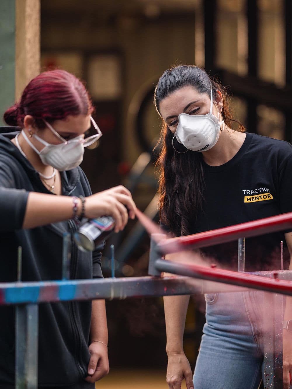 Two people, both wearing white protective masks, are outside spray-painting a bicycle frame. A young woman with red hair is holding the spray can and applying paint, while a woman in a black "Traction Mentor" shirt looks on.