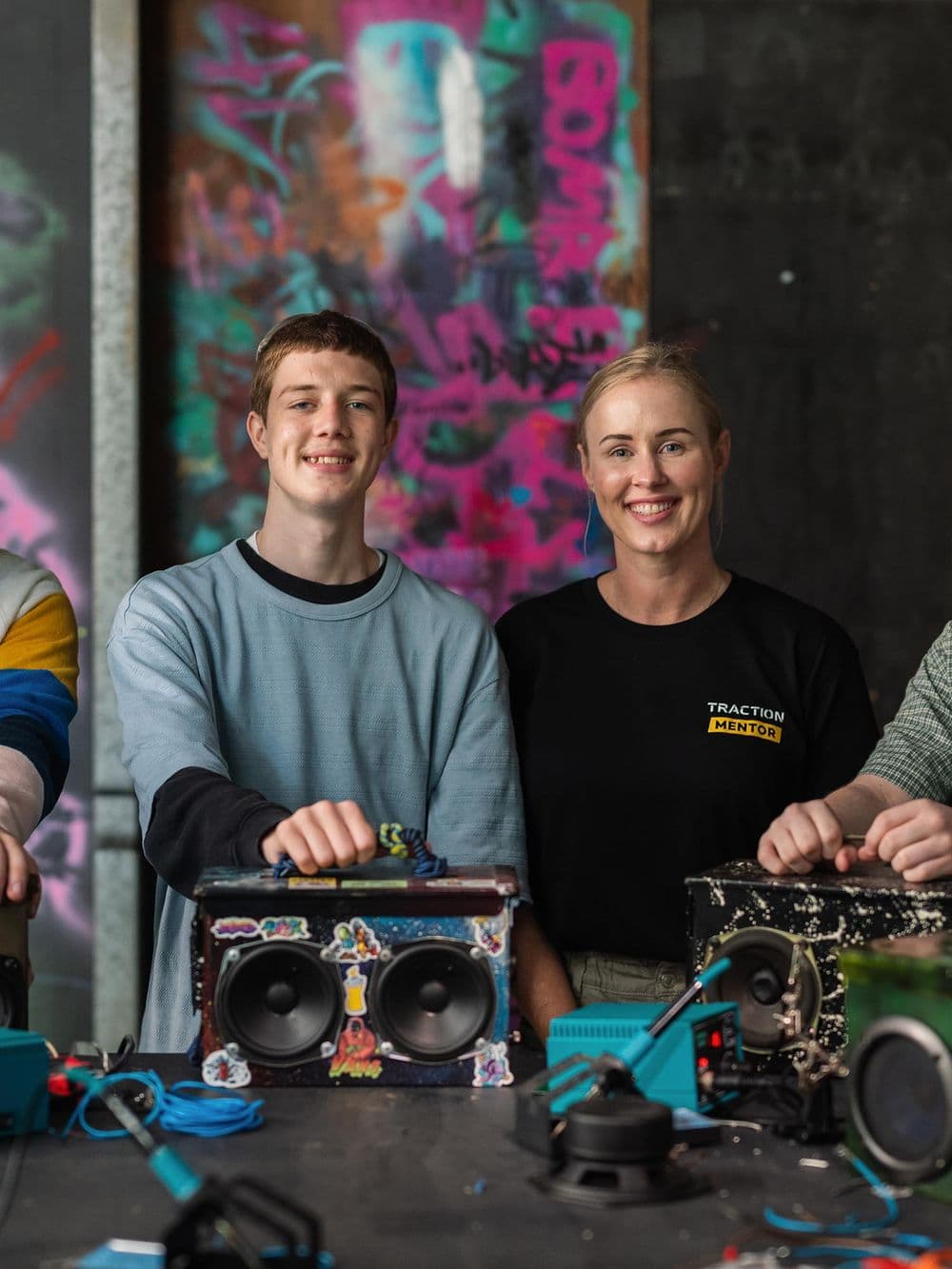 A close-up shot of a young man and a woman in a black "Traction Mentor" shirt, standing behind a table. They are both smiling directly at the camera. The young man has his hands on a colorful, stickered boombox they have built. Graffiti is visible on the wall behind them.