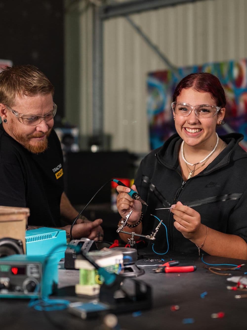 A young woman with red hair and safety glasses is smiling brightly at the camera while practicing soldering a wire with an iron in a workshop. A man with a beard and a black "Traction Mentor" shirt looks on and smiles. Various electronic components and tools are on the table.