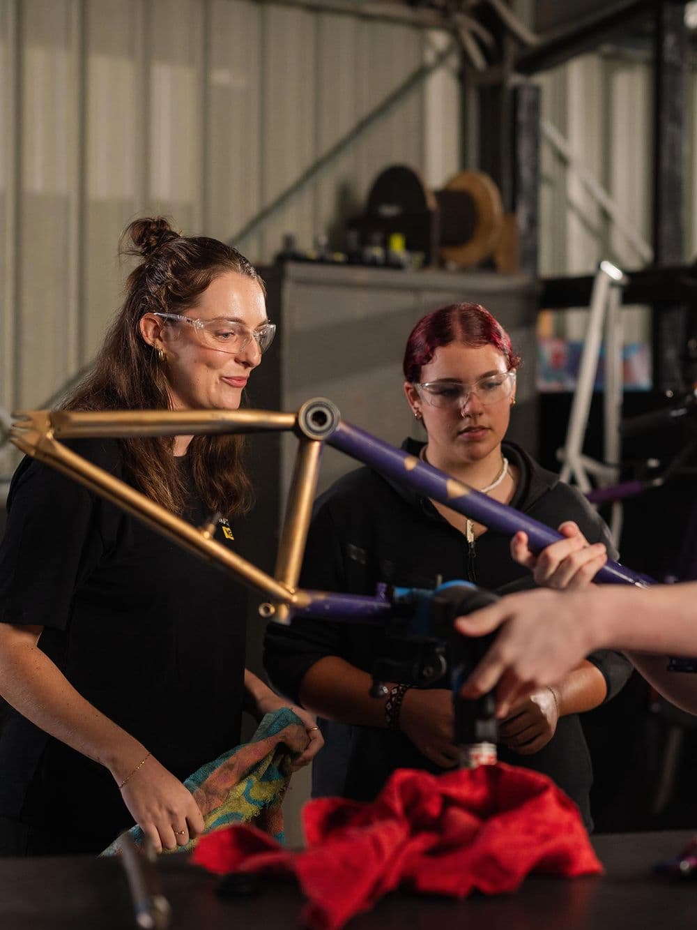 Two young women in safety glasses are working indoors on a bicycle frame. The woman on the left, with her hair in a bun, is smiling and holding a cloth. The woman on the right, with red hair, is holding a purple bicycle frame piece as another person's hands work on it with a tool.